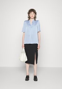 Blusa de cetim azul claro com mangas curtas, combinada com uma saia preta até ao joelho com uma fenda frontal. Segura uma mala branca e usa tamancos pretos.
