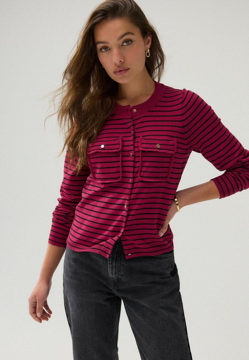 Fuchsie gestreifter Cardigan mit rundem Halsausschnitt, zwei Brusttaschen und goldfarbenen Knöpfen, kombiniert mit dunkelgrauen Jeans.