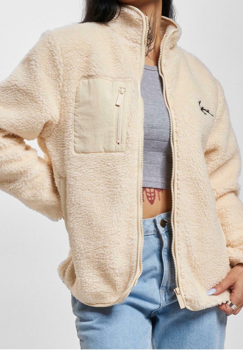 Fleecejacka i ljusbeige med dragkedjeförsedd bröstficka, hög krage och texturerad yta. Har dragkedjestängning och kontrasterande detaljer.