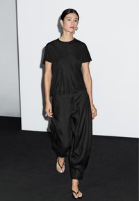 Sort t-shirt med korte ærmer og løstsiddende, sorte, højtaljede bukser. Glat tekstur. Modellen har klare flip-flops på. Minimalistisk design mod en neutral baggrund.