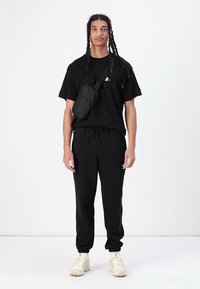 Zwarte t-shirt, zwarte joggers met een trekkoord en witte sneakers. Een kleine zwarte schoudertas wordt op de schouder gedragen.