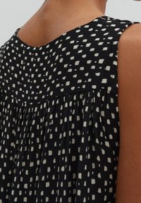 Musta hihaton paita, jossa on rento istuvuus, ja siinä on valkoinen abstrakti pilkkuvaikuutus sekä laskostettu rakenne selän kaula-aukossa.