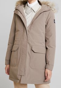 Beige parka, jossa on turkiskaulus, kaksi etutaskua ja vetoketjukiinnitys. Hihansuissa resorit ja pinta on sileä, mattapintainen.