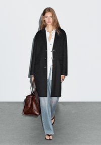 Czarny długi płaszcz o prostym kroju, z zapięciem na guziki, kieszeniami po bokach, noszony na białej koszuli i jasnoniebieskich szerokich dżinsach, w połączeniu z brązową torebką.