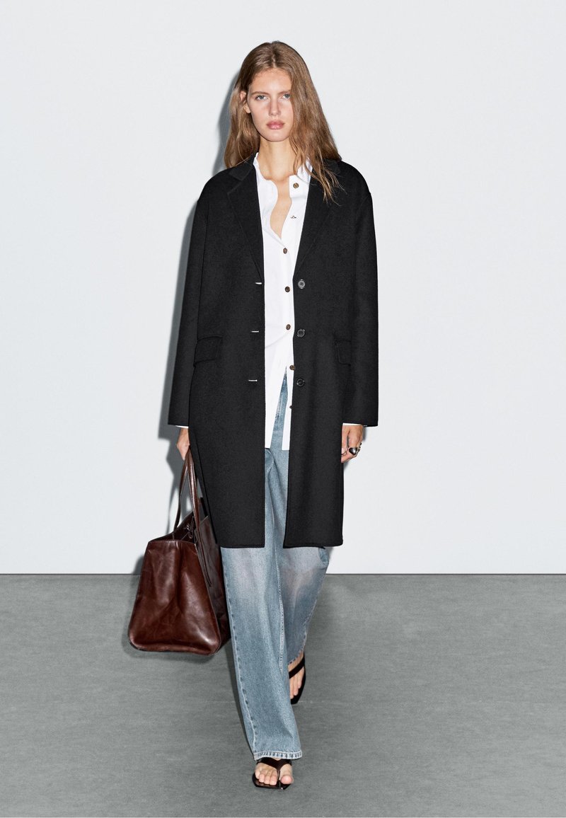 Czarny długi płaszcz o prostym kroju, z zapięciem na guziki, kieszeniami po bokach, noszony na białej koszuli i jasnoniebieskich szerokich dżinsach, w połączeniu z brązową torebką.