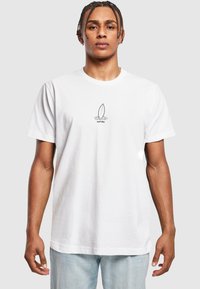 Hvid bomulds T-shirt med rund hals, prydet med et centralt grafisk motiv af et surfboard inden i en cirkel, iført med lyseblå jeans.