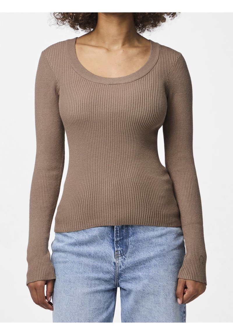 Femme portant un haut marron moulant à manches longues côtelées et un jean bleu clair taille haute sur un fond uni.