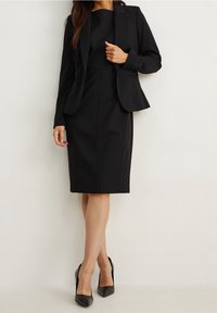 Costume noir sur mesure avec un blazer ajusté et une jupe crayon. Tissu lisse, lignes épurées, accentué par des talons pointus pour un look soigné.