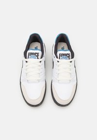 Zapatilla blanca con parte superior de cuero perforado, acentos negros, detalles en azul en la lengüeta y suela exterior de goma negra. Cuenta con cordones planos.