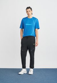 Zils sporta t-krekls ar Reebok logo, baltām kontrastējošām detaļām, kopā ar melnām sporta biksēm ar rāvējslēdzēja kabatām un spīdīgi melnām ielīmēm. Baltas sporta kurpes.