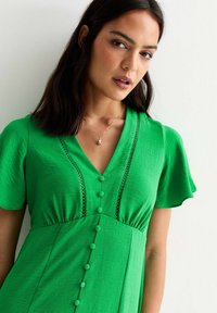 Vestido verde hecho de una tela ligera con mangas cortas, escote en V, adornos de encaje decorativo y botones en la parte delantera.