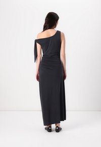 Femme portant une longue robe noire une épaule avec un détail noué sur l'épaule gauche, debout de dos sur un fond blanc uni.