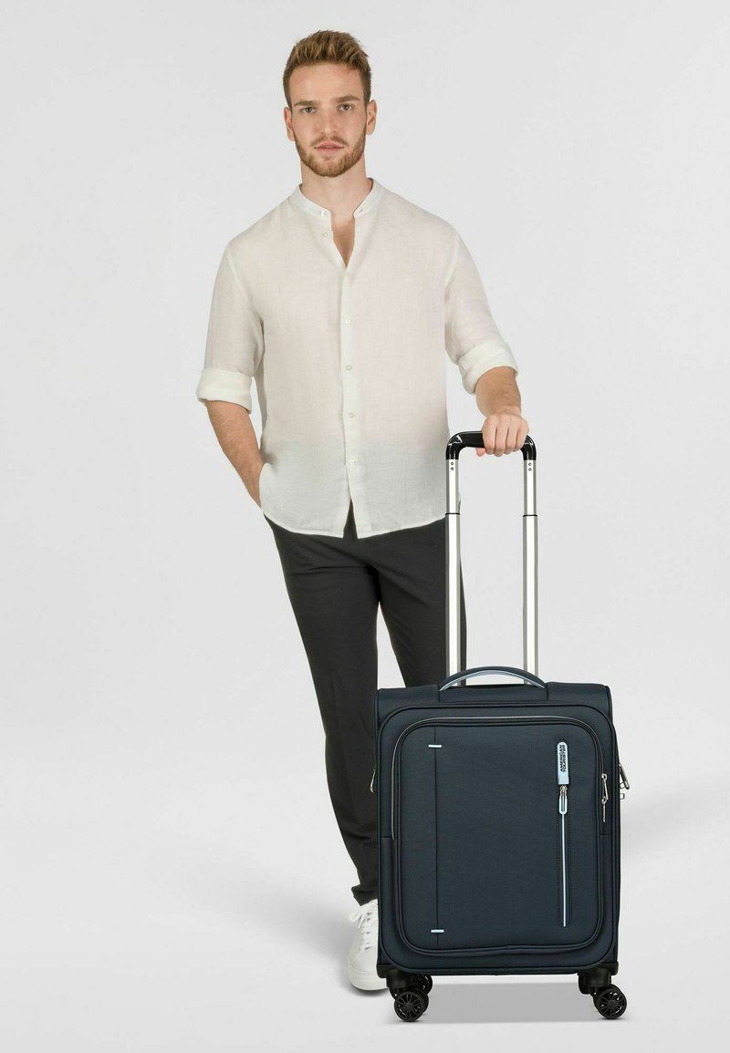 Valigia rigida blu navy con manico retrattile, quattro ruote girevoli e tasche con zip. Superficie texturizzata con accenti blu chiaro.