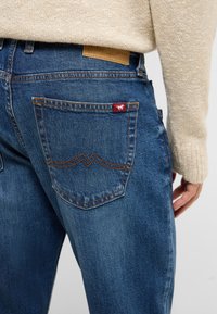 Jean en denim bleu foncé avec coutures orange, présentant un design de poche arrière et une étiquette en cuir de marque au-dessus de la ceinture.