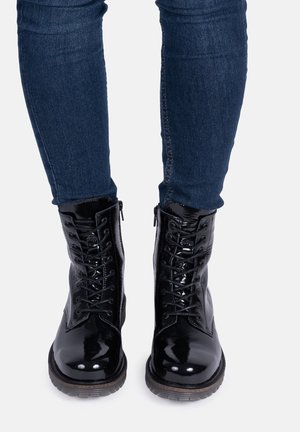 Botas de charol negras con cordones, punta redonda, superficie texturizada y cremallera lateral, combinadas con jeans ajustados de color azul oscuro.
