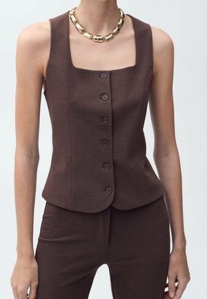 Gilet marron sans manches à boutons, associé à un pantalon marron assorti et un collier chaîne en or massif porté par un mannequin sur un fond uni.