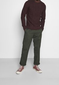 bordeauxfarbener Kabelstrickpullover, olivgrüne Hose und burgunderfarbene Converse-Sneaker. Das Outfit zeichnet sich durch strukturierte Stoffe und eine lässige Passform aus.