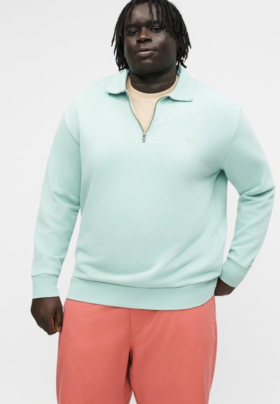 Mladi moški s temno poltjo in dreadlocki nosi svetlo modri pulover z zadrgo do polovice in koralne hlače, stoji pred enobarvnim ozadjem.