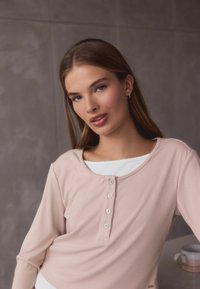 Light pink long-sleeve top with a scoop neckline and five button placket. Soft fabric, fitted design, layered over a white shirt.