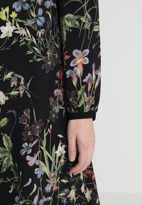 Svart blommig klänning med en variation av färgglada blommönster. Ärmarna är långa med en samlad manschettdetailj. Mjuk tygtextur.