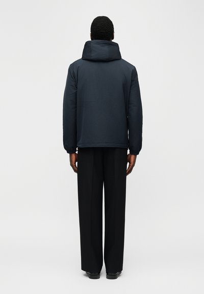 Άνδρας με την πλάτη, φορά σκοτεινό μπουφάν με κουκούλα, μαύρο παντελόνι φαρδιάς γραμμής και μαύρα παπούτσια, μπροστά σε ένα απλό φωτεινό φόντο.