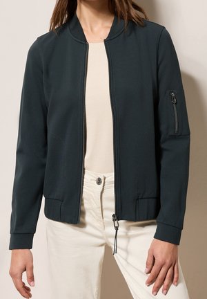 Femme portant une veste bomber vert foncé zippée avec des poches latérales et une poche zippée sur la manche gauche, associée à un pantalon beige et un haut.
