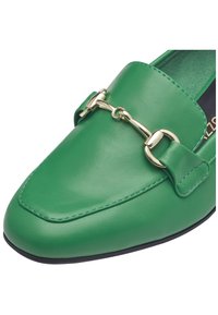 Mocasines de cuero verde con punta redonda, detalle de hardware dorado y una textura suave con una suela negra contrastante.
