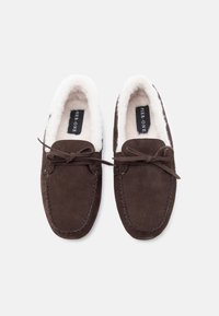 Καφέ σουέτ παντόφλες μοκασίνια με λευκή γούνα εσωτερικά και λεπτομέρειες από φιόγκο, θέα από ψηλά σε άσπρο φόντο.