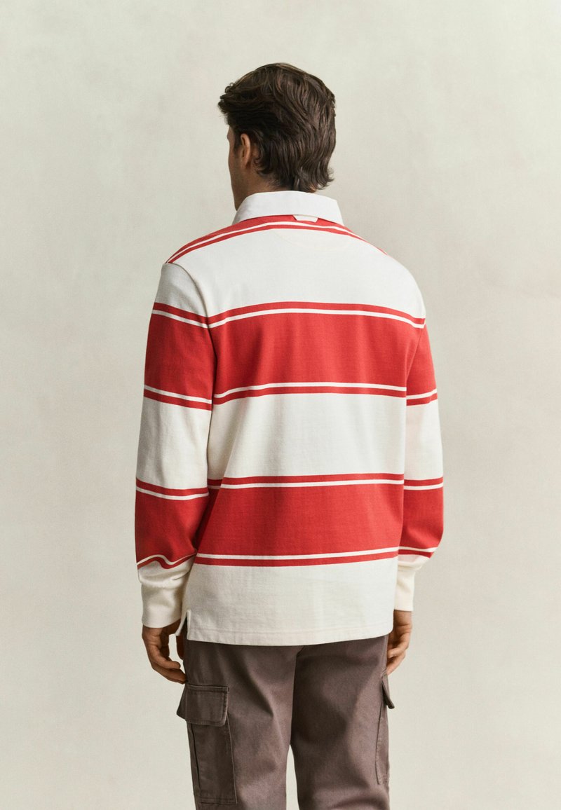 Mann trägt ein langärmliges Rugbyshirt mit breiten roten und weißen horizontalen Streifen, steht mit dem Rücken zur Kamera, kombiniert mit braunen Cargohosen.