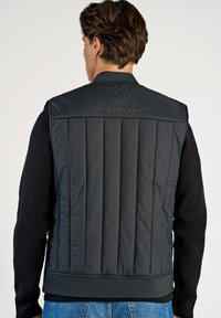 Svart vadderad väst med hög krage, utrustad med horisontell sömnad och ribbad nederkant. Varumärket "LINDBERGH" är präglat på baksidan.