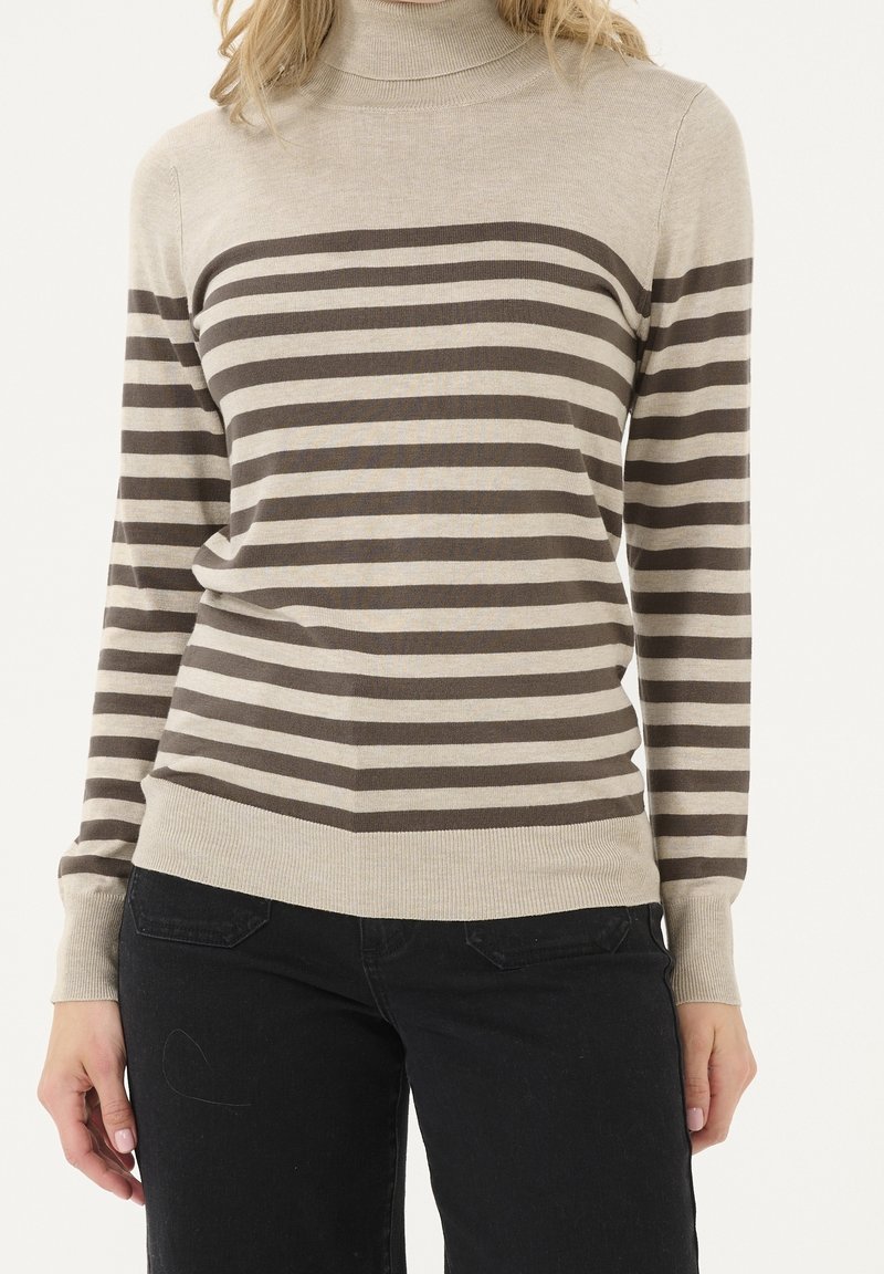 Femme portant un pull à col roulé beige avec des rayures horizontales marron foncé, associé à un pantalon noir, posant devant un fond uni.