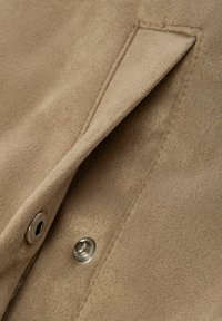 Tessuto scamosciato beige con un primo piano di una patta di tasca, caratterizzata da un bottone in argento e dettagli di cucitura, che evidenziano una consistenza morbida.