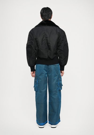 Chaqueta bomber negra con cuello de piel, combinada con pantalones cargo azules de corte holgado que tienen bolsillos laterales y un dobladillo en contraste.