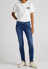 Camiseta corta blanca con la impresión en negro de "Pepe Jeans", jeans skinny de denim azul y zapatillas beige. Conjunto casual simple.