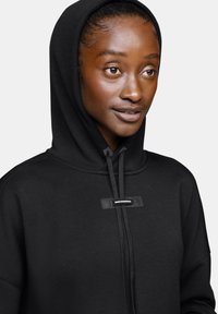 Haut à capuche noir avec cordon de serrage, comportant une étiquette en caoutchouc sur le devant. Fabriqué en tissu doux avec une texture lisse et une coupe oversize.