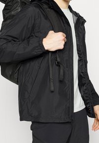 En person klädd i en svart zip-up jacka och bär en svart ryggsäck över ena axeln, iförd svarta byxor och en ljusgrå skjorta.