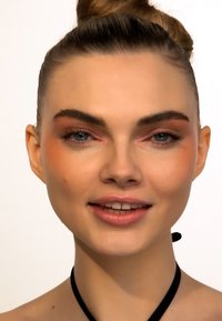 Nærbilde av en models ansikt med glatt hud, ferskenfarget roughe, rosa lepper og gråblå øyne. Håret er stylet i en toppknute; hun har på seg en svart choker.