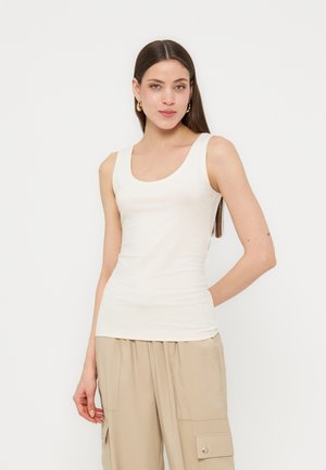 Młoda kobieta o długich brązowych włosach ubrana w biały bezrękawnik i beżowe spodnie typu cargo, pozująca na jednolitym białym tle.