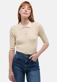 Polo shirt beige à côtes avec manches courtes, patte de boutonnage à trois boutons, associé à un jean bleu en denim taille haute. Texture lisse, coupe décontractée.