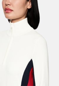 Weißer Zip-Pullover mit geripptem Kragen und Stoffpanelen in Marineblau und Rot an den Ärmeln, die ein modernes Design und eine interessante Textur betonen.