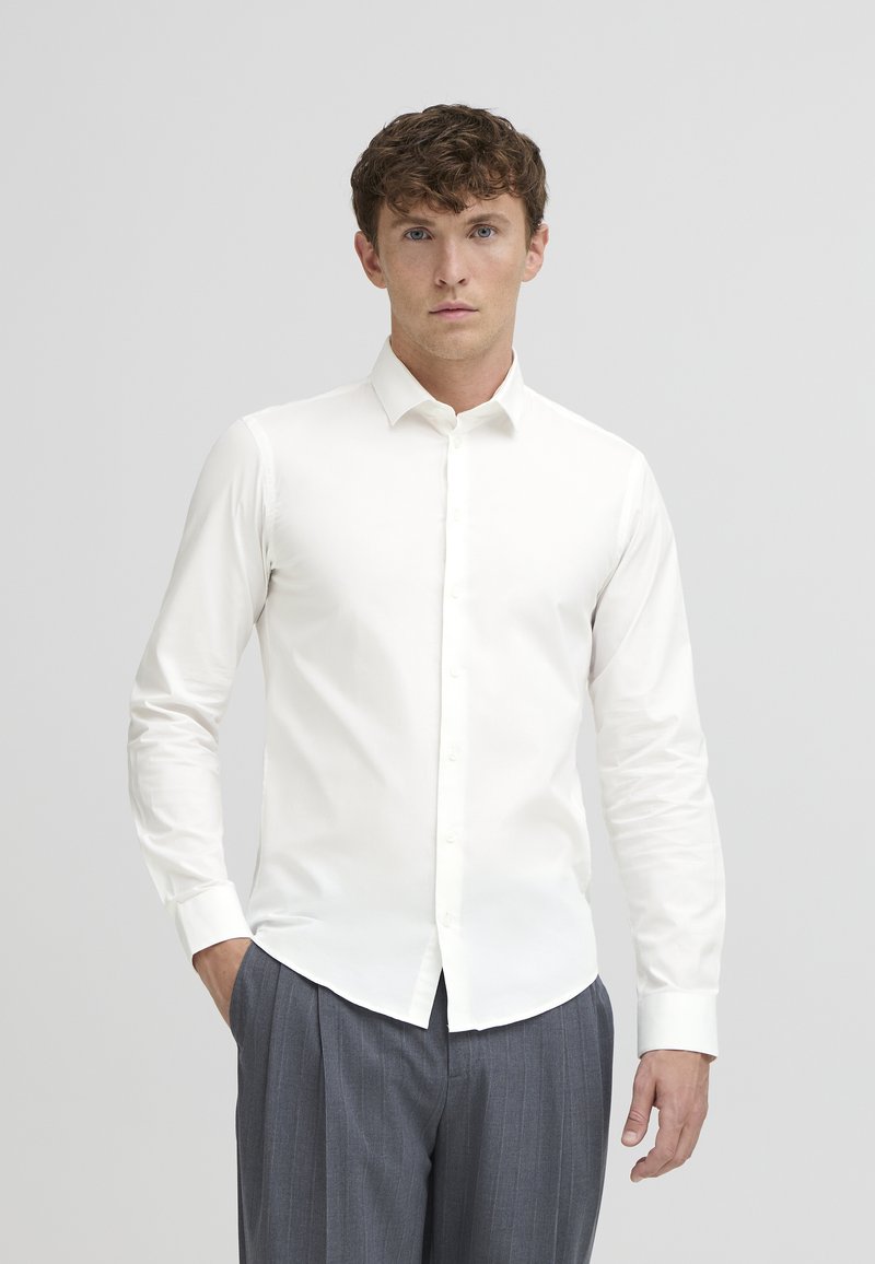 White, long-sleeve dress shirt with a pointed collar and button front. Smooth fabric, fitted design, paired with grey pleated trousers.