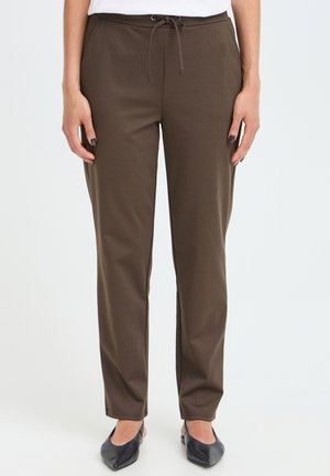 Femme portant un pantalon droit marron foncé avec taille à cordon et des ballerines noires pointues, debout devant un fond uni.