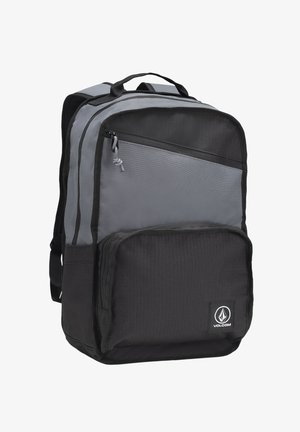 Mochila gris y negra hecha de un tejido duradero, que cuenta con múltiples compartimentos con cremallera, un asa superior y un parche con el logo en la parte frontal.