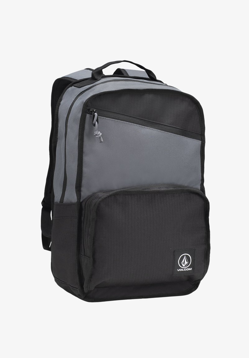 Mochila gris y negra hecha de un tejido duradero, que cuenta con múltiples compartimentos con cremallera, un asa superior y un parche con el logo en la parte frontal.