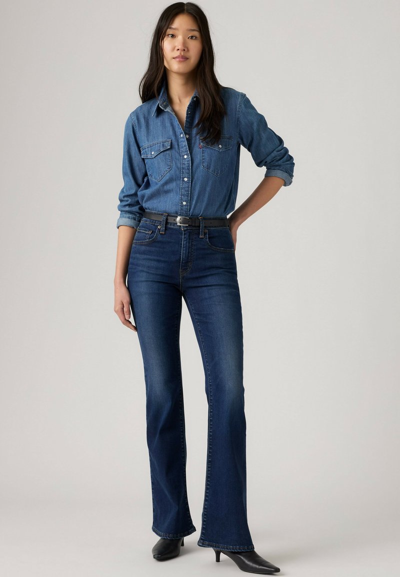Femme debout portant une chemise en jean bleue à manches retroussées, un jean évasé bleu foncé avec une ceinture noire, et des bottes noires à bout pointu.