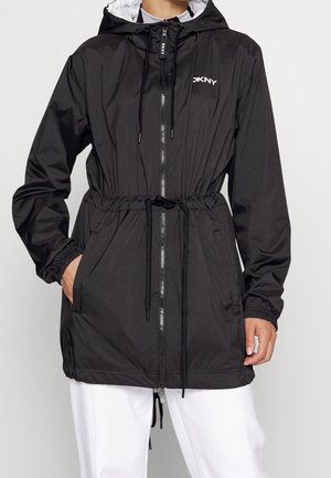 Person trägt eine schwarze DKNY Regenjacke mit Kapuze, Frontreißverschluss, Kordelzug in der Taille, elastischen Bündchen und Taschen, kombiniert mit weißen Hosen.
