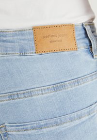 Ljusblå denimjeans med en texturerad yta, iögonfallande med en brun läderlapp märkt "perfekta jeans" och kontrasterande sömnadsdetaljer.