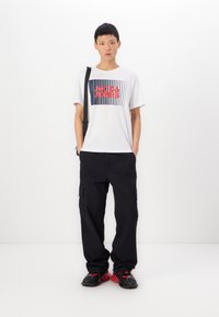 Weißes T-Shirt mit grafischem Druck in Marineblau und Rot, kombiniert mit schwarzen Cargo-Hosen mit Seitentaschen und schwarzen Sneakers mit roten Akzenten.