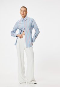 Camisa a rayas azules con líneas verticales rojas, de mangas largas, con botón en la parte delantera y un lado abierto. Combinada con pantalones anchos blancos.