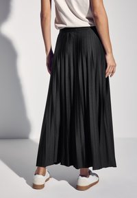 Zwarte plissé maxirok met een elastische tailleband, gecombineerd met witte sneakers. De stof heeft een gladde textuur en een vloeiende ontwerpen.